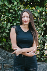 A brunette female with long hair wearing a blank black t-shirt i while standing on a concrete wall background on a street. Empty space for text or design.