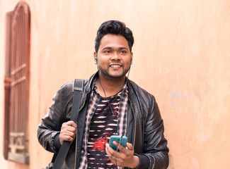 Young indian man holding mobile phone with earphones is walking  confident - Happy asian male using smartphone smiling and looking ahead - Concept of lifestyle  and tecnology with pink wall copy space