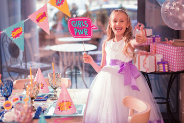 Excited super girl. Emotional long haired girl wearing white dress and putting thumb up while expecting the party