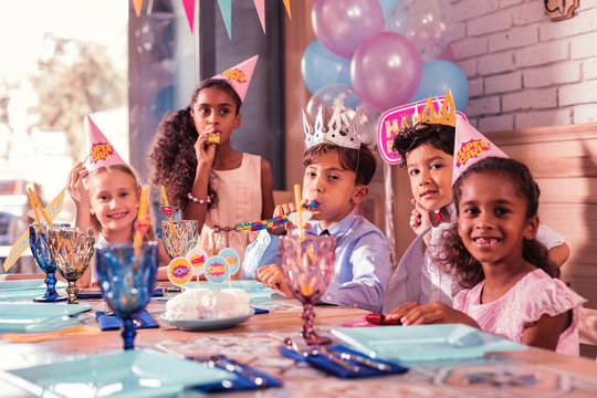Five children. Positive relaxed pretty kids sitting together at the birthday party and blowing lovely party horns