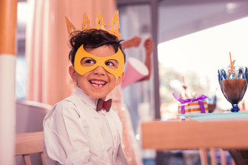 Ready for carnival. Cheerful smiling little boy sitting at the carnival and wearing paper mask with...