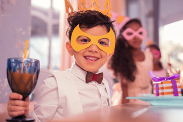 Masquerade. Cheerful smiling boy feeling happy while being at the masquerade and drinking juice
