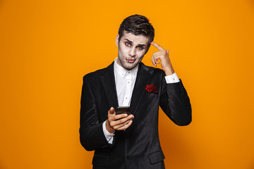 Photo of handsome dead man on halloween wearing classical suit and creepy makeup holding mobile phone, isolated over yellow background