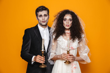 Photo of spooky zombie couple bridegroom and bride wearing outfit and halloween makeup holding candles, isolated over yellow background