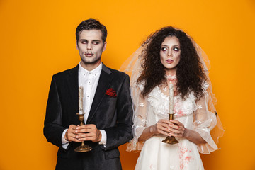 Photo of attractive zombie couple bridegroom and bride wearing outfit and halloween makeup holding candles, isolated over yellow background