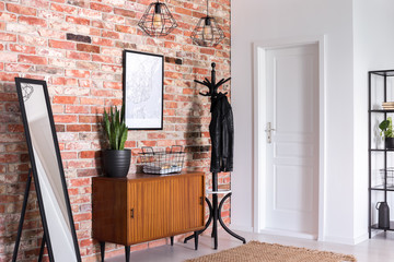 Mirror next to wooden cabinet in entrance hall interior with white door and poster on red brick wall. Real photo