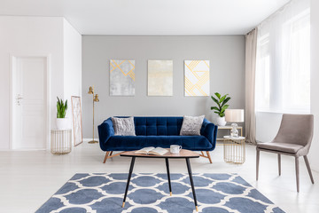 Open space living room interior with modern furniture of a navy blue settee, a beige armchair, a coffee table and other objects in gold color. Real photo.