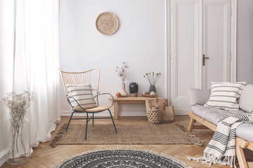 Armchair and sofa with patterned pillows in white flat interior with plants and round rug. Real...