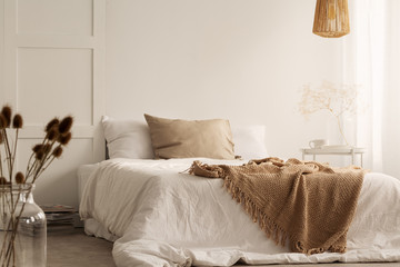 Flowers and lamp in white natural bedroom interior with blanket and pillows on bed. Real photo - obrazy, fototapety, plakaty