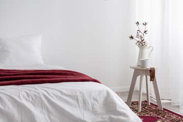Flowers on table next to white bed with red blanket in minimal bedroom interior. Real photo