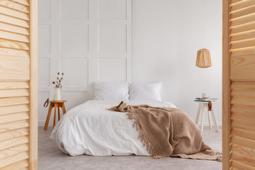 Wooden door and lamp in white bedroom interior with blanket on bed and flowers on table. Real photo