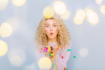 Happy blond curly hipster girl enjoying and celebrating on a grey background. Blows up multicolored confetti. Photo taken through the garland light. Leisure, winter, holidays, people concept.