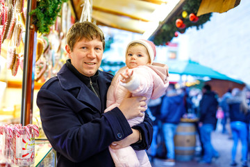 Middle aged father holding baby daughter near sweet stand with gingerbread and nuts. Happy family on Christmas market in Germany. Cute girl eating cookie called Lebkuchen. Celebration xmas holiday.