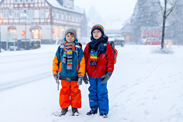 Two little kids boys of elementary class walking to school during snowfall. Happy children having fun and playing with first snow. Siblings ans friends with backpack in colorful winter clothes.
