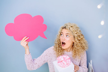 Sale concept. Surprised blonde wopman with pink paper cloud like think do not know what to buy for present or gift. Female sits indoor over grey wall background. Winter holiday concept.