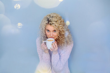 Attractive blond curly woman with a cup of coffee indoor over grey wall background. Photo taken through the garland light. Leisure, winter, holidays, people concept.