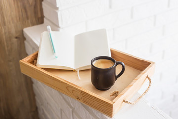 A cup of coffee and an open notebook on a wooden tray in the light interior of the apartment. Cozy home
