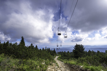 Onboard a ski lift going up a mountain at summertime with people. Cloudy and forest in Rila...