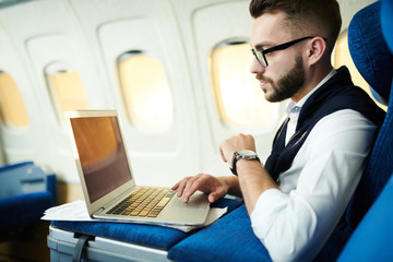 Fototapeta premium Side view portrait of handsome businessman using laptop while working in plane during long first class flight, copy space