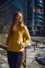 A young European woman, traveling, with long blond hair, wearing a yellow jacket, yellow sunglasses walking down the city center street, street shooting. Even light.
