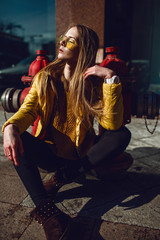 A young European woman, traveling, with long blond hair, wearing a yellow jacket, yellow sunglasses walking down the city center street, street shooting. Even light.