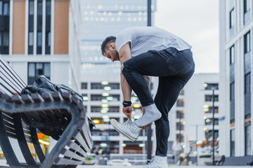 Distant plan of man putting on his white sneakers and standing next bench.