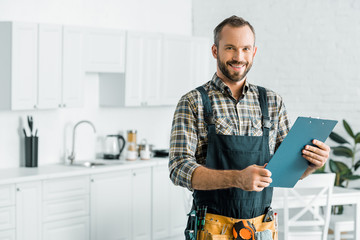 smiling handsome plumber holding clipboard and looking at camera in kitchen - obrazy, fototapety, plakaty