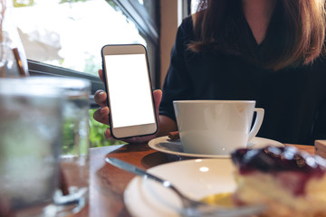Mockup image of a woman holding and showing white mobile phone with blank screen with coffee and cake on the table in cafe