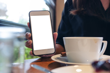 Mockup image of a woman holding and showing white mobile phone with blank screen with coffee and cake on the table in cafe