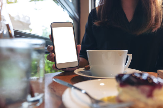 Mockup image of a woman holding and showing white mobile phone with blank screen with coffee and cake on the table in cafe