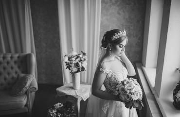 A beautiful bride with a bouquet in her hands is standing by the window. Blonde in a lace dress....