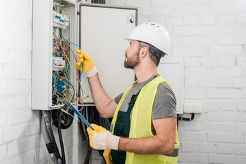 side view of electrician holding clipboard and checking wires in electrical box in corridor