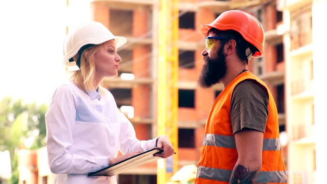 The head of construction discusses details of work with workers on the construction site. A woman in a white helmet communicates with a bearded man in a protective vest on the construction site.