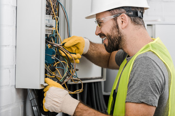 side view of smiling handsome electrician repairing electrical box with pliers in corridor