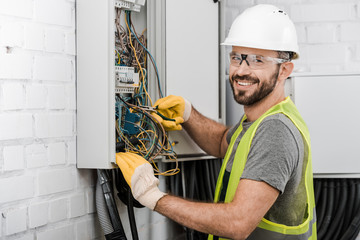 Fototapeta smiling handsome electrician repairing electrical box with pliers in corridor and looking at camera obraz