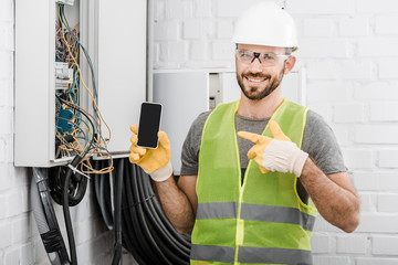 smiling handsome electrician pointing on smartphone with blank screen near electrical box in...