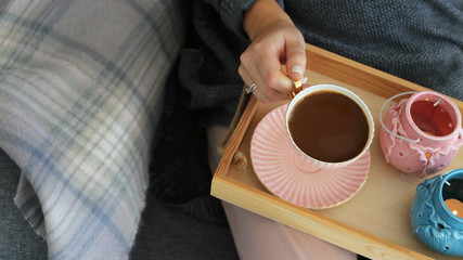 A cup of coffee with milk in female hands on a wooden tray with pink and turquoise candlesticks on a gray cozy sofa with a checkered powder rug. Interior and home cosiness concept. Top view