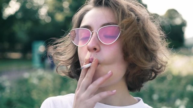 Young woman with short fair hair wearing pink sunglasses and t shirt smoking cigarette and drinking coffee in summer park. Concept of addiction and bohemian lifestyle. Slider slow motion portrait shot