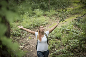 Young woman with backpack and raised up arms while walking through the forest.