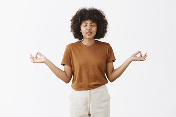 Indoor shot of calm and pacient stylish urban female student with afro hairstyle and dark skin closing eyes smiling carefree spreading hands aside in zen gesture meditating and relieving from stress