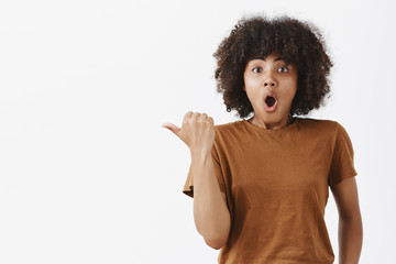 Uplifted excited cute dark-skinned girl with curly hair in brown trendy t-shirt pointing left with...