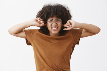 Fototapeta na wymiar Cute good-looking female nerd with afro hairstyle in transparent prescribed glasses frowning and wrinkling nose clenching teeth from dislike and discomfort covering ears not hear noisy bothering sound