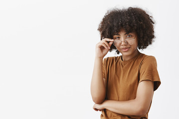 Studio shot of smart african american female genius with afro hairstyle in trendy brown t-shirt looking from under glasses with knowing look being suspicious smirking as if having great plan