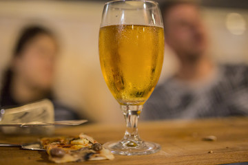 image with detail of a slice of pizza with a glass of beer and two people in the background
