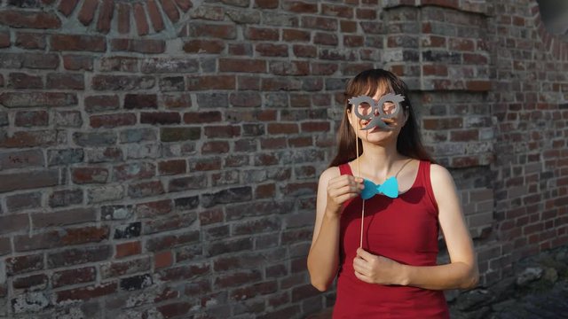 A young happy woman holds a paper black mustache and a a blue bow tie on a stick while dancing next to a brick wall.