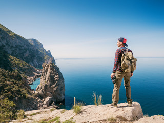 A young man stands on the edge of the mountain and looks at the sea. Guy tourist with binoculars enjoys the seaside.