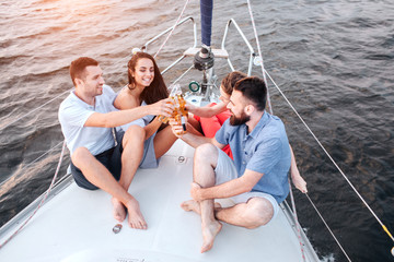 Two couples sitting on bow of yacht and cheers with beer. They smile. Men keeps legs crossed. All...