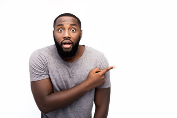 Headshot of attractive African man looking at camera with amazed expression eyes and full disbelief, pointing his index finger at blank copy space wall, shocked with content