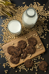 top view of the chocolate chip cookies on the wooden tray, a glass of soy milk, a pile of soybeans, a bottle of soy milk with dried wheat bouquet and the napery.