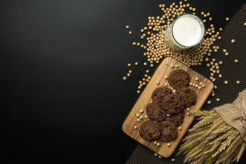 Fototapeta na wymiar top view of the chocolate chip cookies on the wooden tray, a glass of soy milk, a pile of soybeans and dried wheat bouquet on the napery, black background